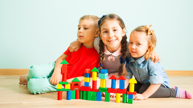 Drei Kinder stapeln auf dem Boden sitzend bunte Holzbausteine.