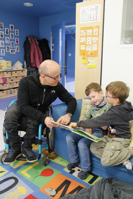 Mann im Rollstuhl zeigt zwei Jungs, die auf der Couch sitzen, etwas in einem Kinderbuch.